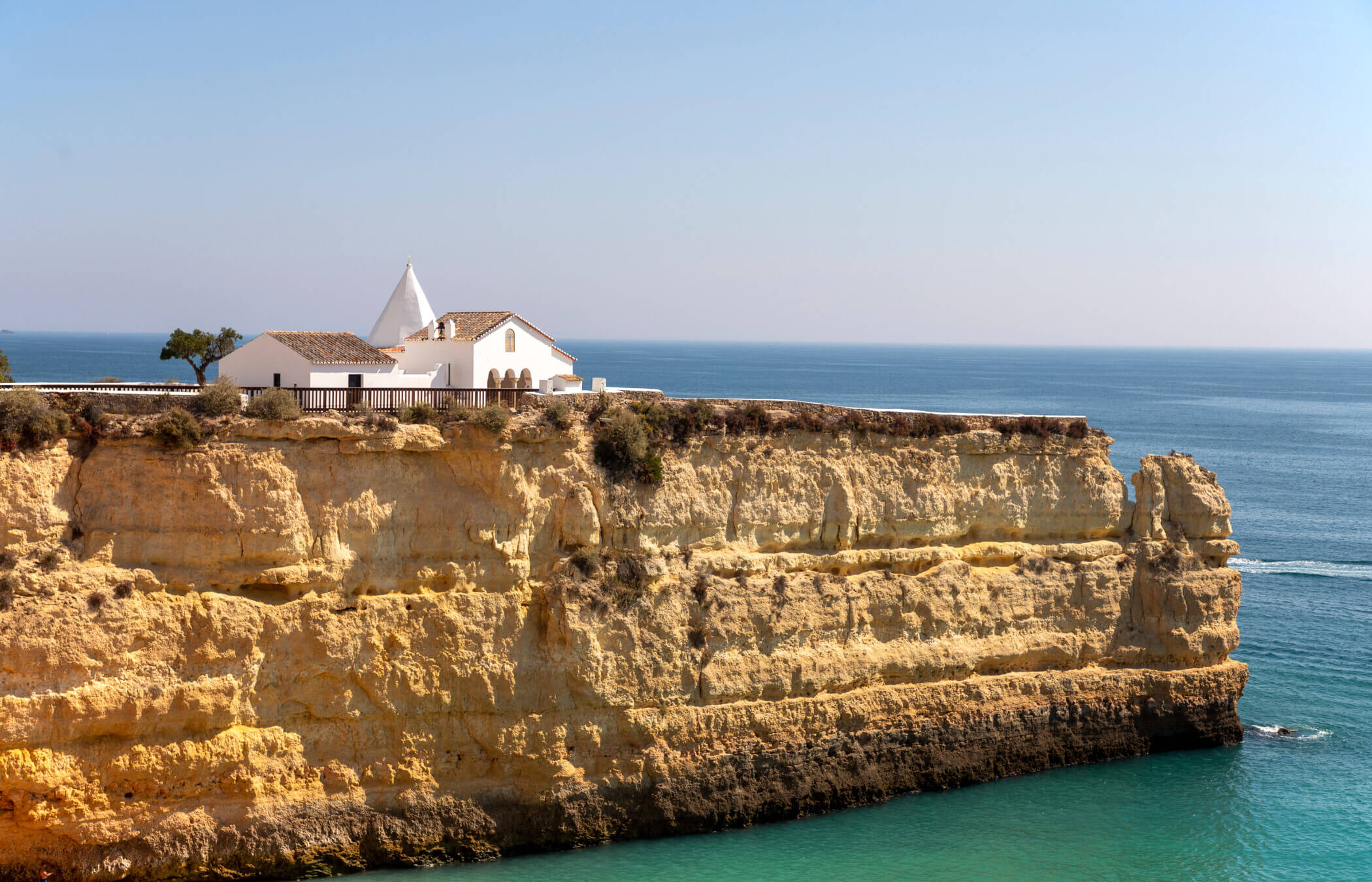 Eine weiße Kirche thront auf einer goldenen Klippe, umgeben vom blauen Meer. Das ist Nossa Senhora da Rocha (Unsere Liebe Frau vom Felsen) an der Algarve in Portugal.
