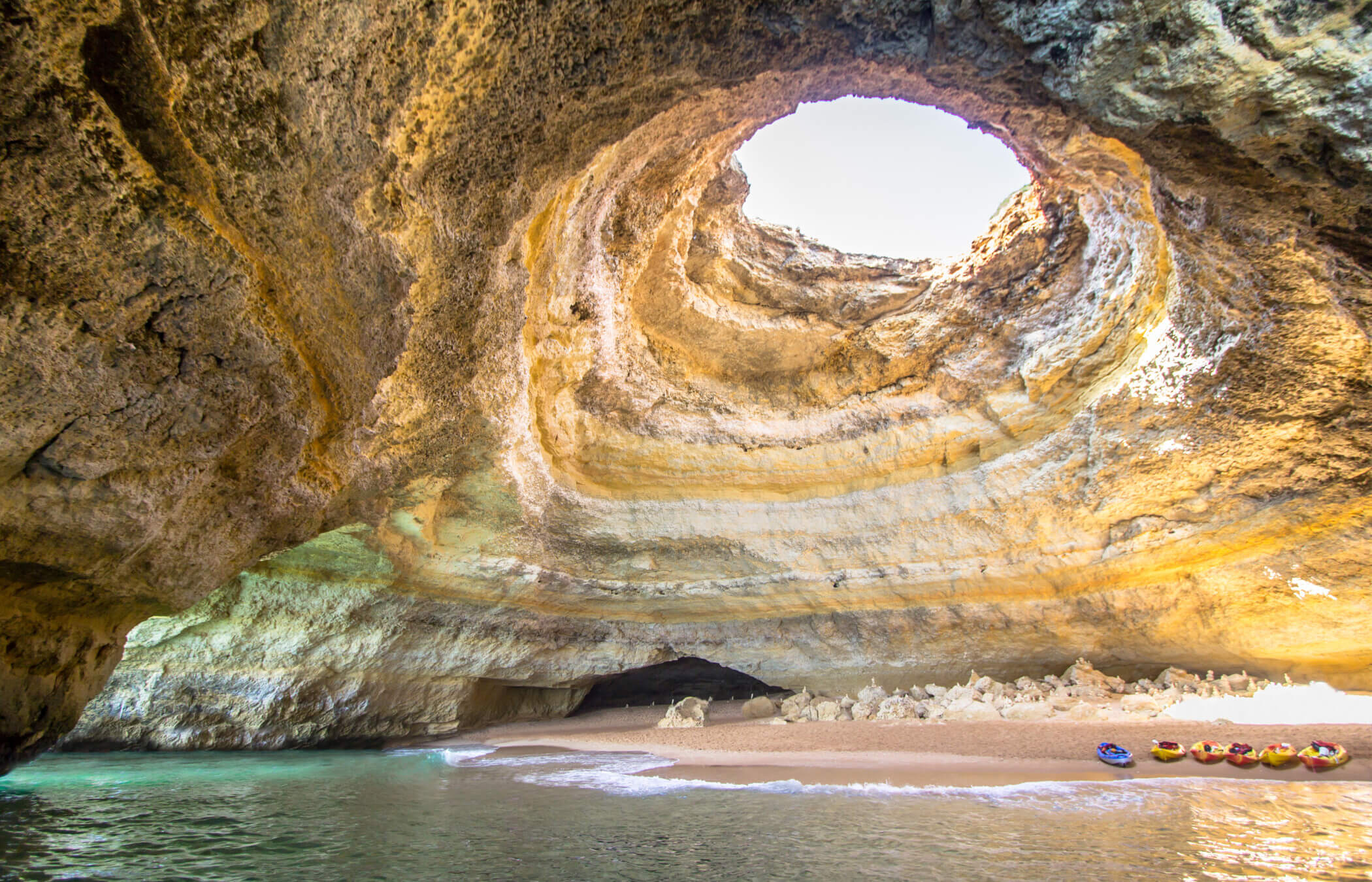 Einige Kajaks liegen an einem kleinen Strand, während das Wasser an das Ufer einer großen Höhle heranrollt, deren Inneres durch ein großes „Oberlicht“ erhellt wird. Das ist die Benagil-Höhle in Portugal.