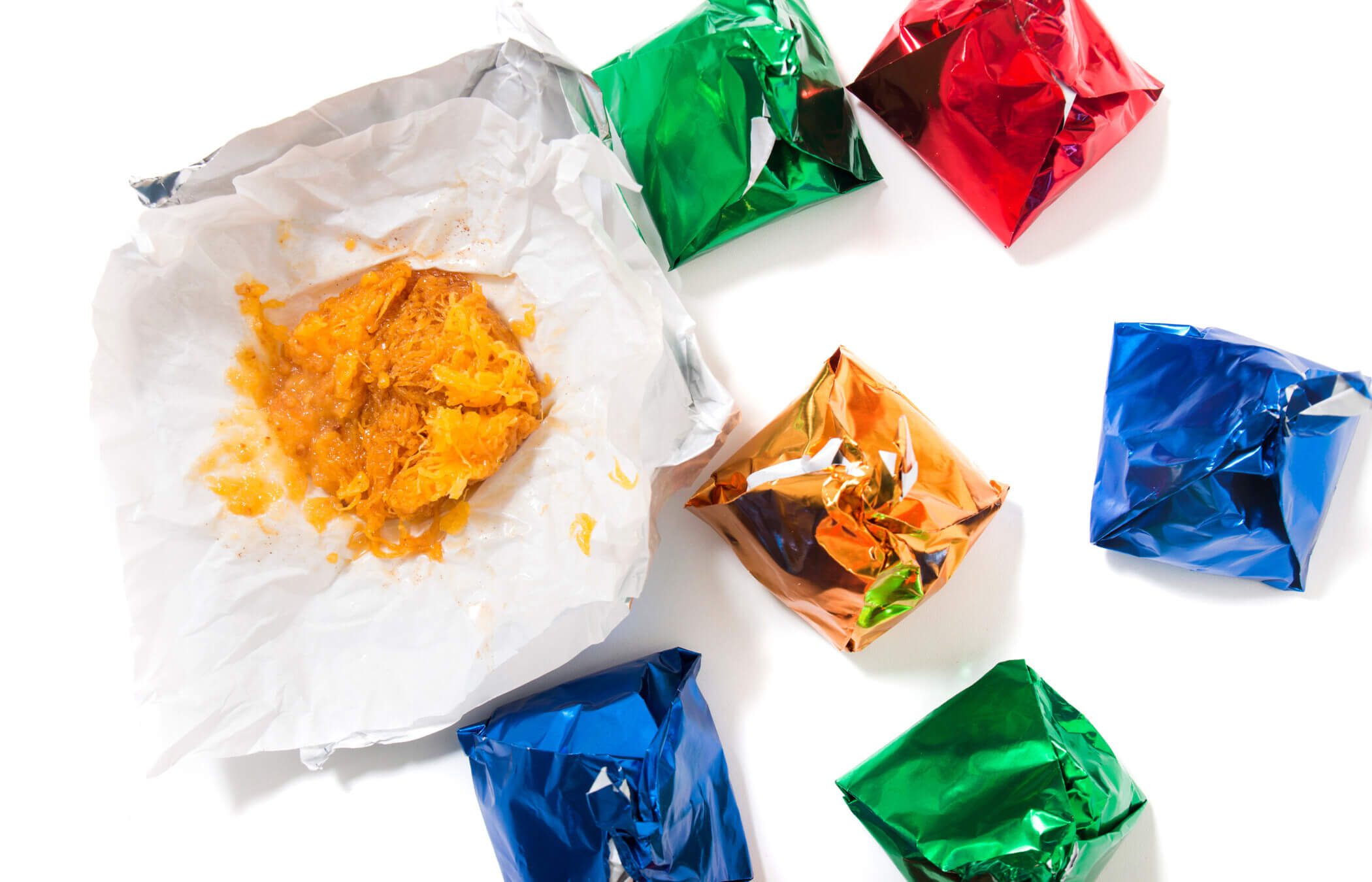 Zeven 'Dom Rodrigo'-desserts staan op een witte achtergrond. Ze zijn allemaal verpakt in kleurrijke folie – groen, blauw, rood en oranje – op één na, die openligt en een papperige oranje massa laat zien.