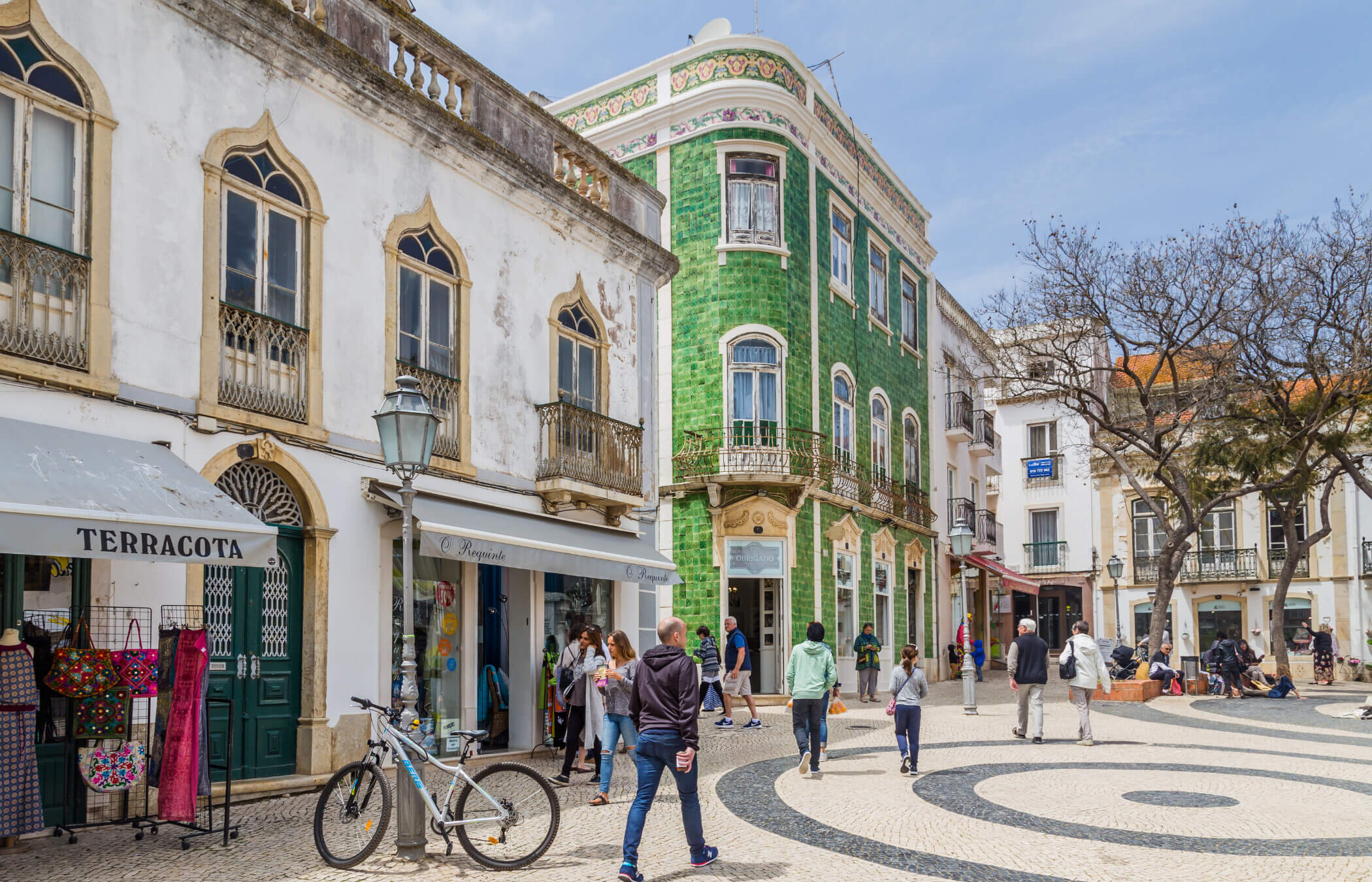 Ein lebhafter Stadtplatz mit Fliesen in wirbelnden Mustern auf dem Boden, an dem ein auffälliges grünes Gebäude steht, das mit aufwendigen ornamentalen Verzierungen geschmückt ist. Jacaranda-Bäume ragen über Parkbänke empor, während Menschen dort sitzen und spazieren gehen und die Atmosphäre genießen.