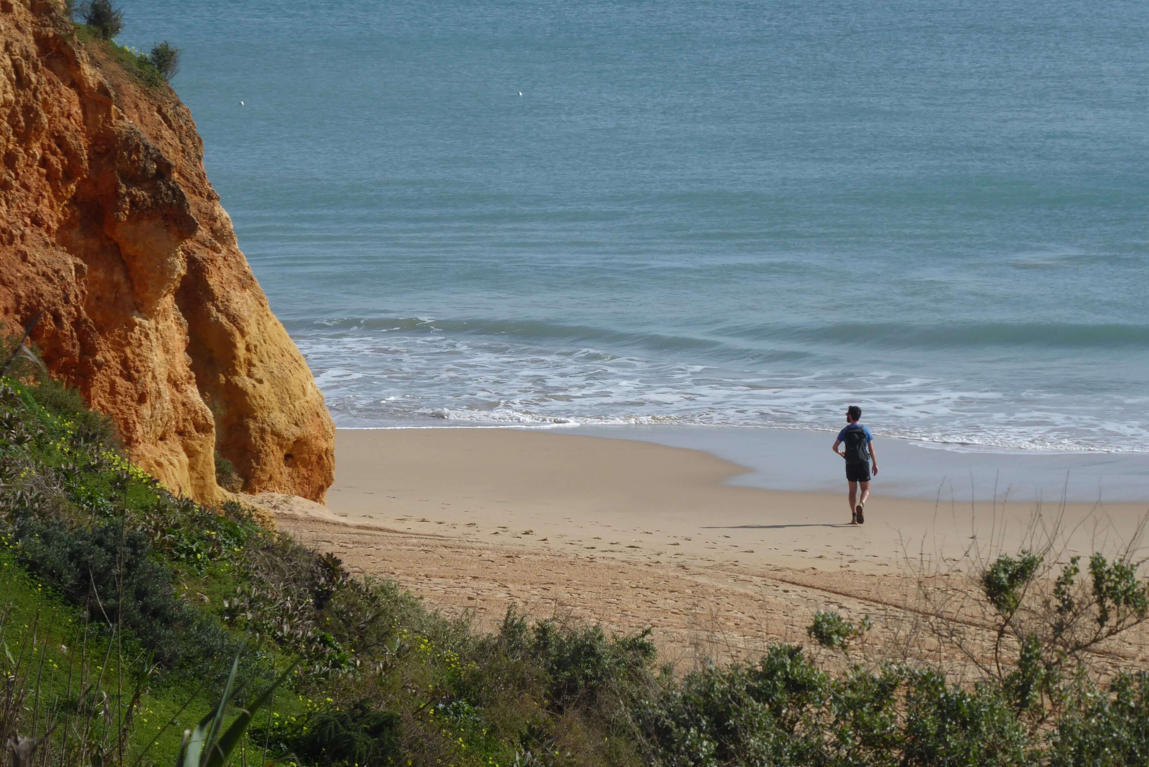 Ein einsamer Wanderer an einem Sandstrand in Alvor bei Portimão blickt auf die Klippen und geht auf den Atlantik zu 