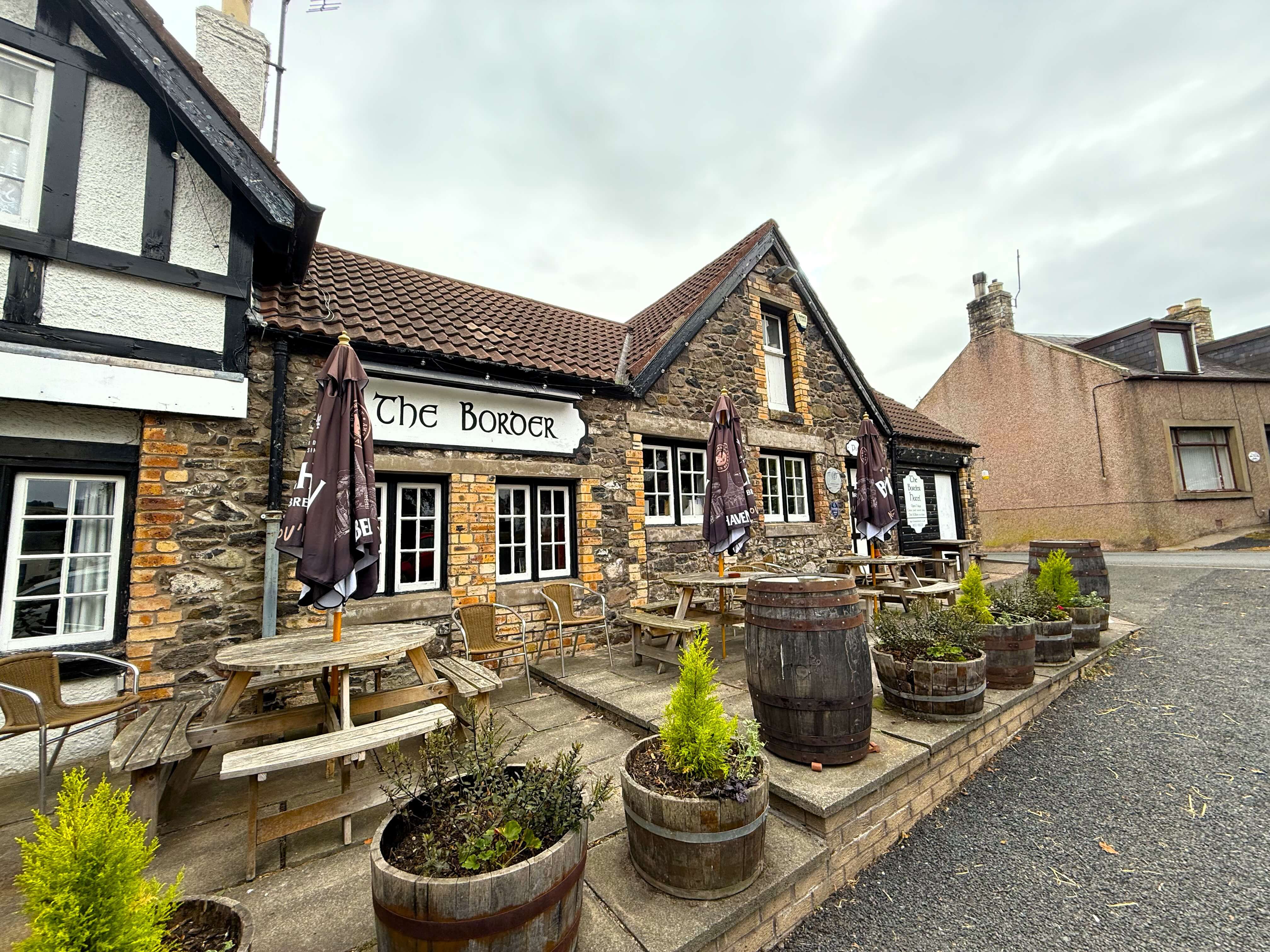 A quaint old pub with charming brickwork. The sign reads 'The Border'. Outside are some traditional wooden beer garden tables with umbrellas, and the area is decorated with old barrels, some of which have been repurposed as planters containing small trees and green plants.