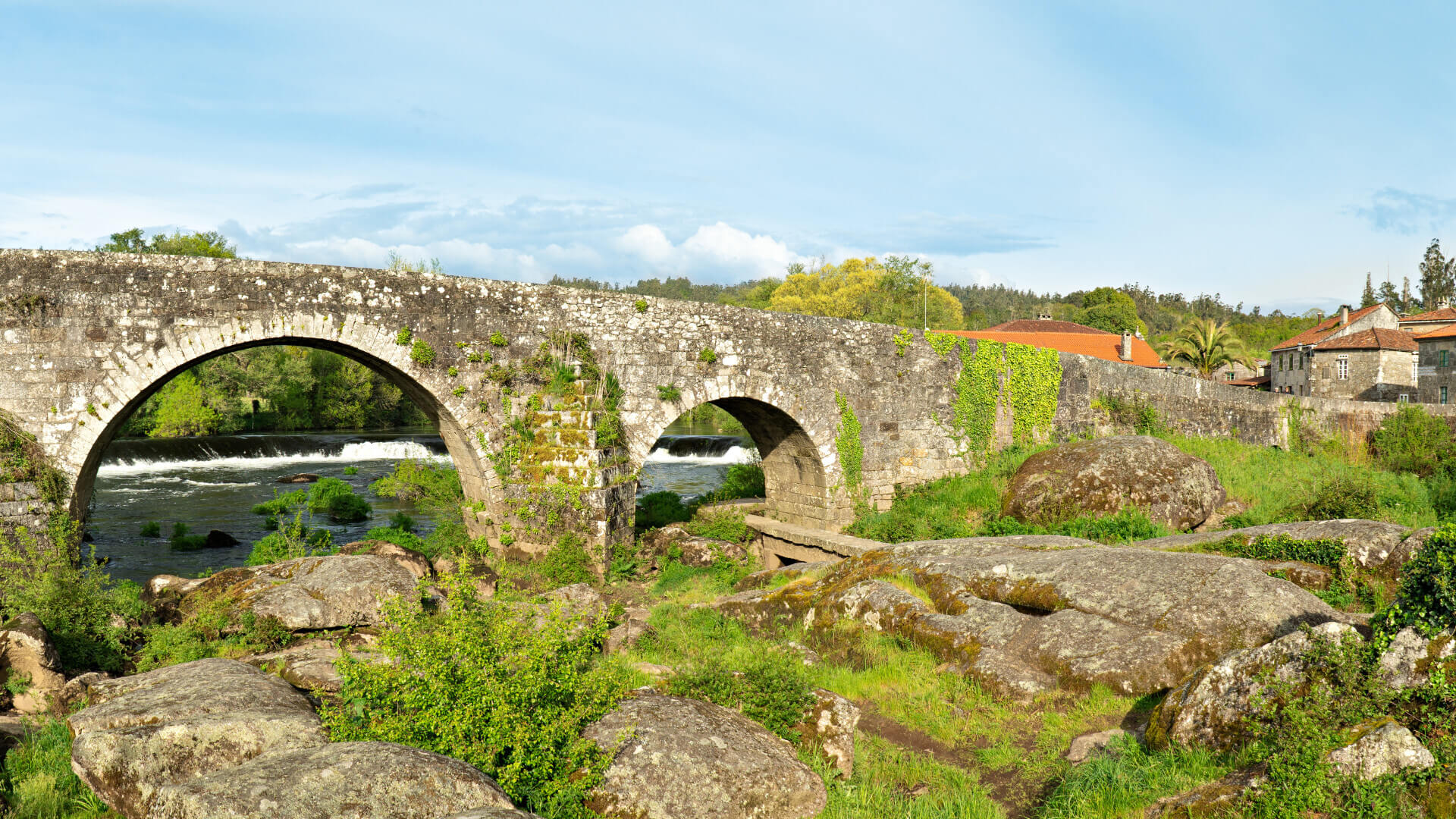 Un ancien pont en pierre à plusieurs arches. À droite se trouve un ensemble de vieux bâtiments en pierre aux toits rouges.