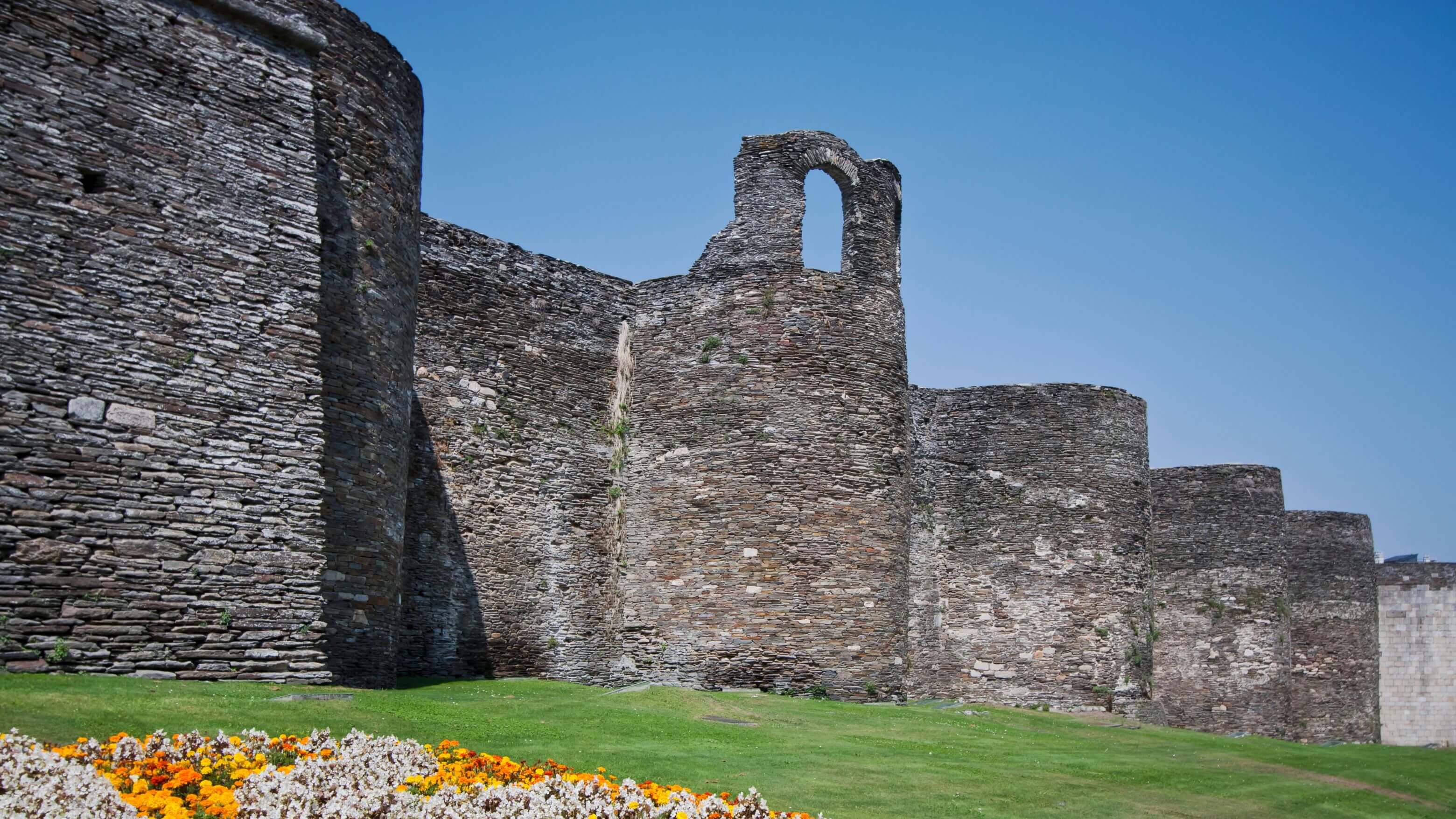 Les imposants remparts romains de Lugo, datant du IIIe siècle, sont construits à partir de milliers de fines pierres d'ardoise empilées horizontalement, créant ainsi une texture rayée caractéristique sur leur surface gris-brun. Ces fortifications imposantes s'élèvent en plusieurs niveaux, avec une tour cylindrique à gauche et une arcade partiellement en ruines au centre. Les remparts s'étendent en courbe au loin, révélant leur échelle gigantesque. Au premier plan, une pelouse soigneusement entretenue et un parterre de fleurs orange et blanches contrastent avec la maçonnerie ancienne patinée par les intempéries, sur fond de ciel bleu limpide. Ce sont les seules murailles romaines intactes qui subsistent au monde, marquant le point de départ du chemin de pèlerinage du Camino Primitivo.