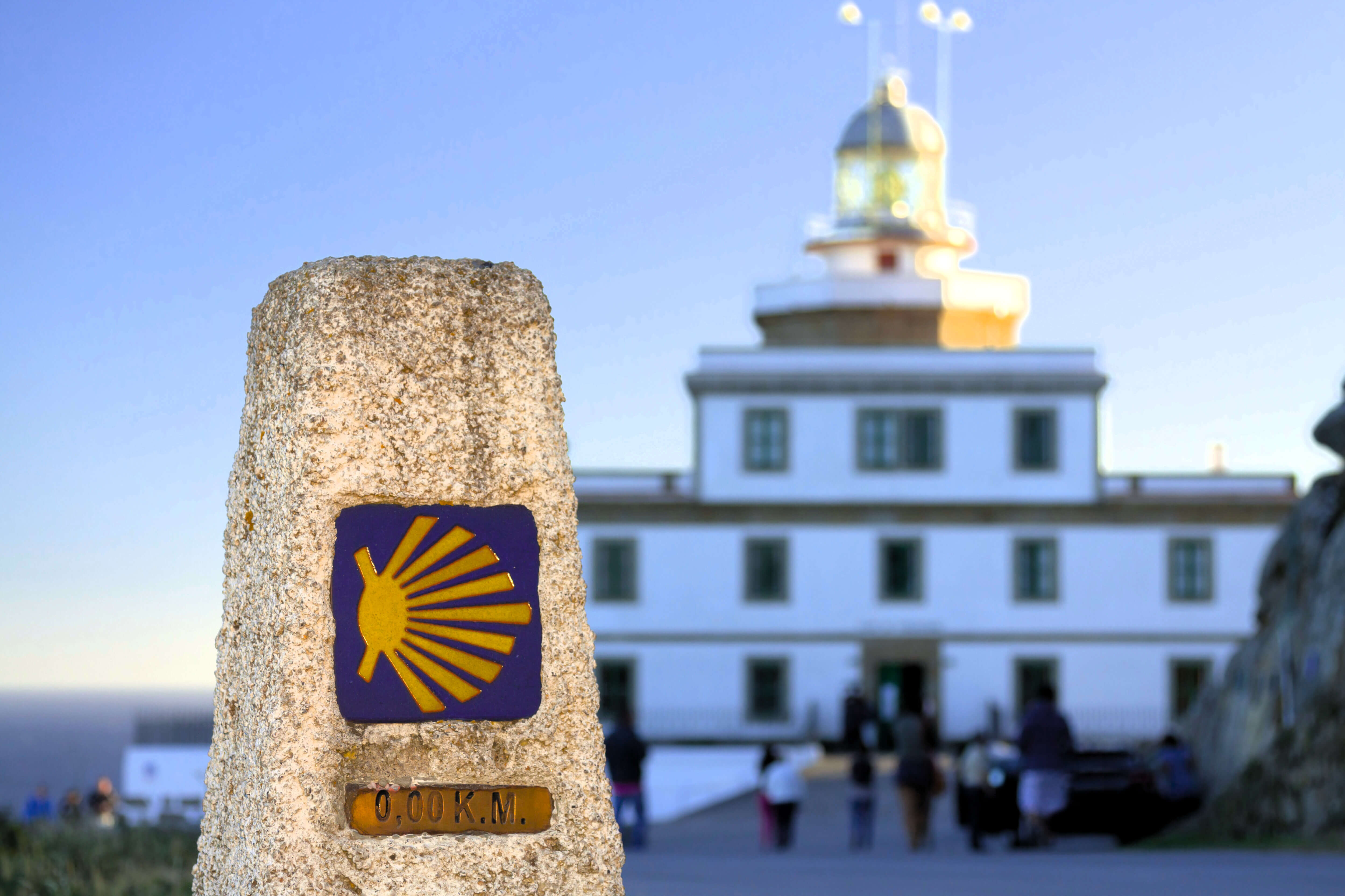 Au premier plan : une borne en pierre ornée d'une coquille Saint-Jacques. À l'arrière-plan : un grand phare. Le phare est situé au sommet d'un bâtiment rectangulaire de trois étages.