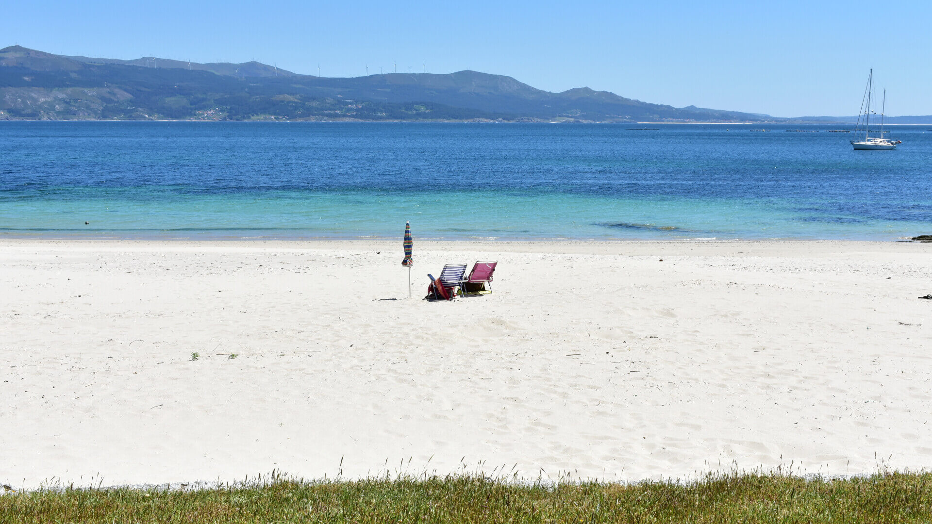 Two foldable chairs and a folded in parasol sit alone on a white sandy beach. A sail boat can be seen in the water. In the background we can see rugged ground beyond the water lined with turbines