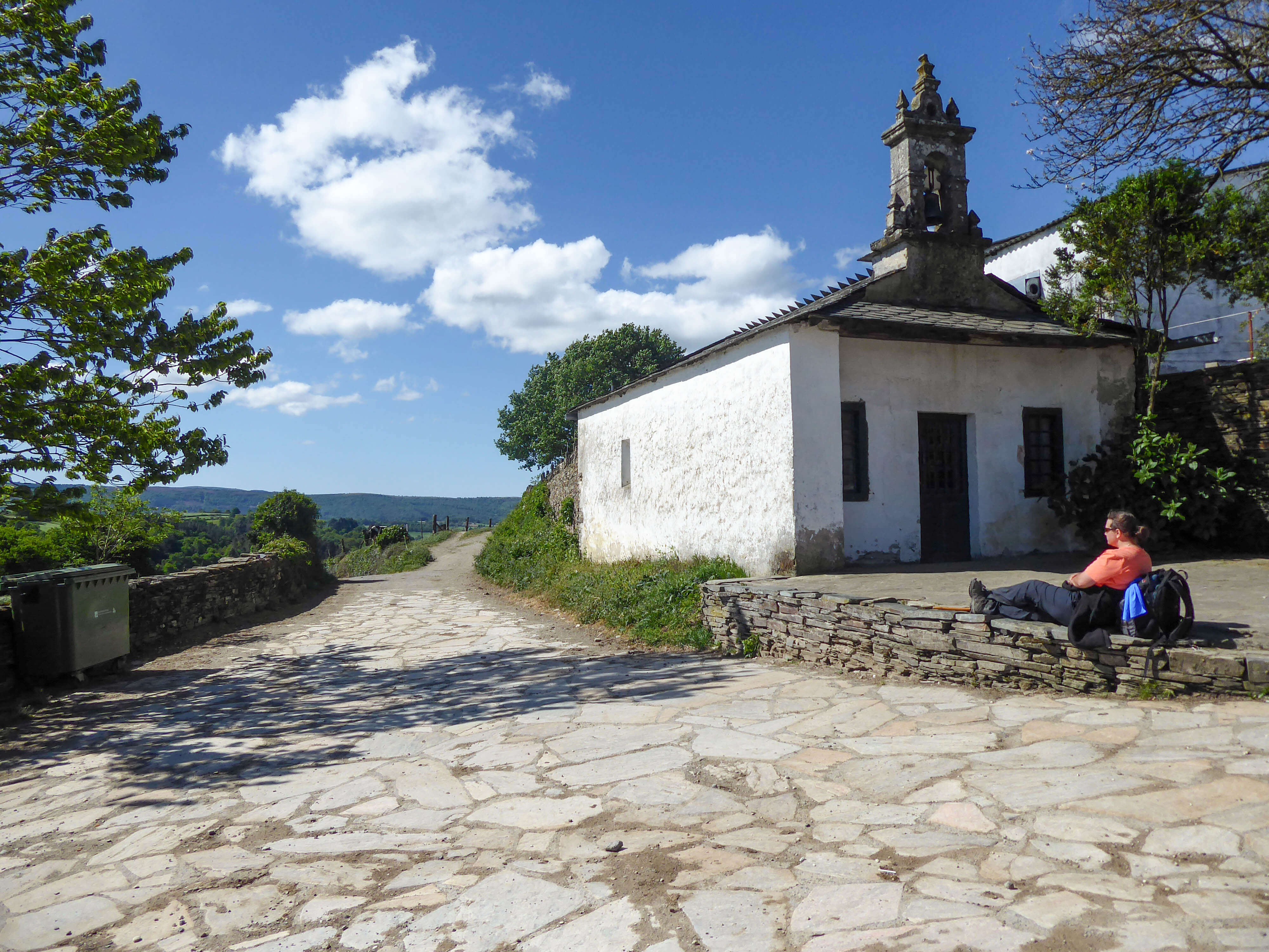 Un randonneur repose ses pieds contre un mur, adossé à son sac à dos, près d'une petite église blanche, le long d'un chemin pavé entouré d'herbe et d'arbres.