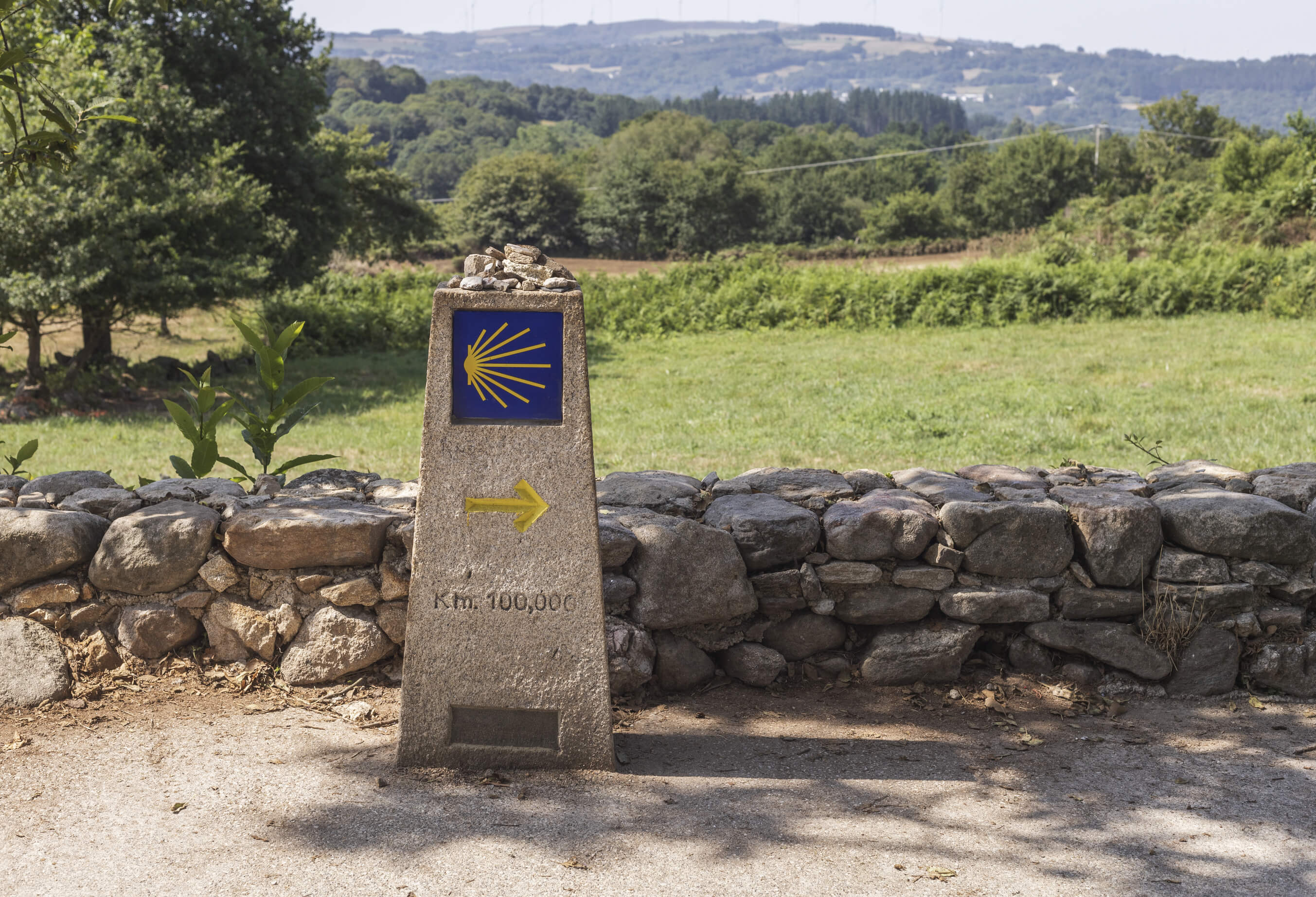 Au premier plan : un repère orné d'une coquille Saint-Jacques. À l'arrière-plan : une campagne verdoyante.
