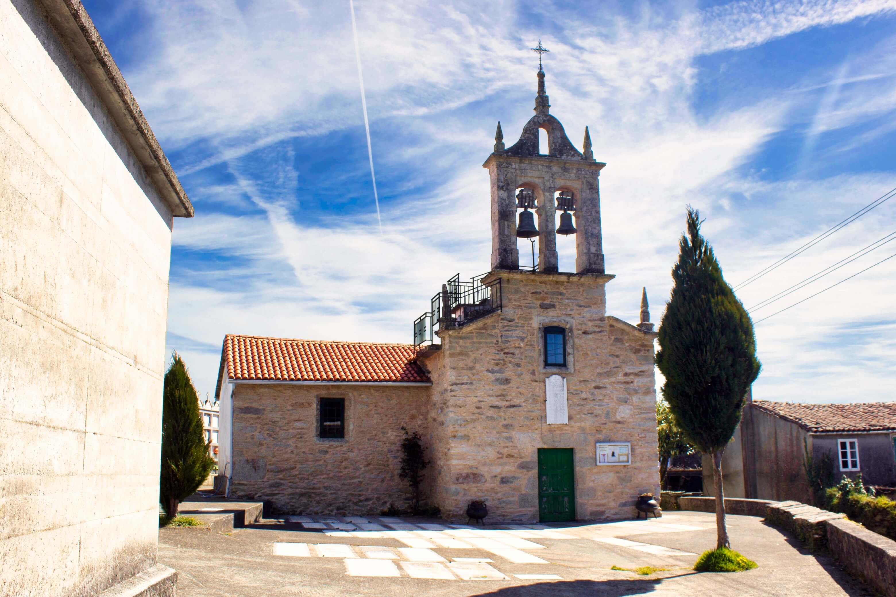 Petite église de village en pierre, dotée d'un clocher abritant deux cloches et surmonté d'une croix. L'église, avec sa porte verte et son toit de tuiles rouges, se dresse sur une place pavée où se trouve un arbre élancé, sous un ciel d'un bleu éclatant parsemé de nuages vaporeux, à Arzúa.
