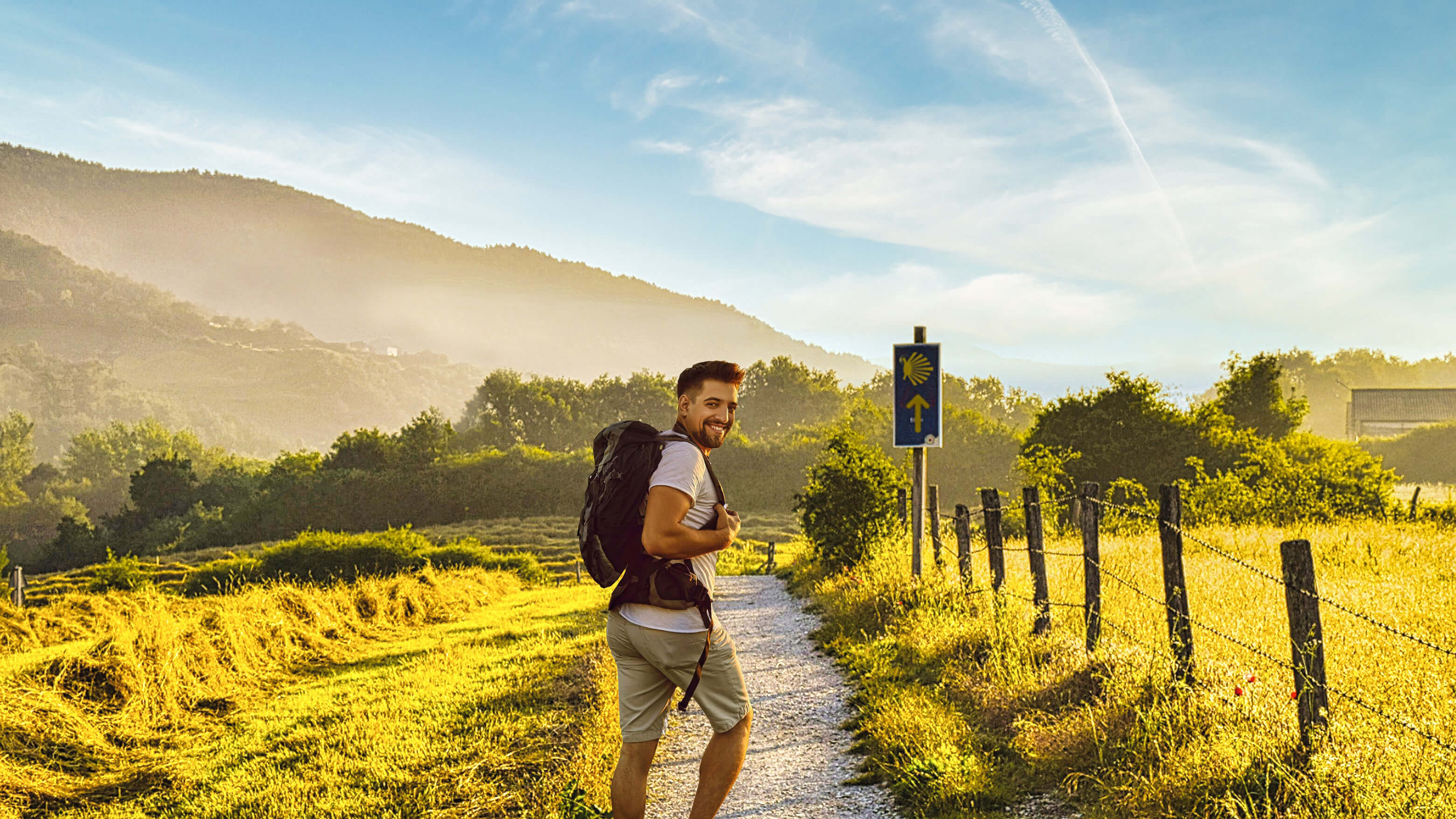 Un jeune pèlerin souriant parcourt le Camino Primitivo sur un chemin de gravier à travers la Galice rurale, à l'heure dorée. Il porte un sac à dos noir, un t-shirt blanc et un short beige, et jette un regard par-dessus son épaule vers l'appareil photo avec une expression joyeuse. Le soleil de fin d'après-midi baigne le paysage d'une lumière ambrée et chaleureuse, illuminant les champs d'herbe dorée de part et d'autre du chemin, séparés à droite par un poteau en bois et une clôture métallique. Au premier plan se dresse un panneau indicateur bleu du Camino, arborant le symbole emblématique de la coquille Saint-Jacques jaune et une flèche pointant vers le haut. Derrière le pèlerin, des arbres verts et une végétation dense bordent le chemin, tandis que des collines boisées s’élèvent dans le fond brumeux, sous un ciel bleu pâle strié de fins nuages. La scène capture la beauté paisible et le sentiment d’accomplissement qui définissent l’expérience du pèlerinage sur le Camino.