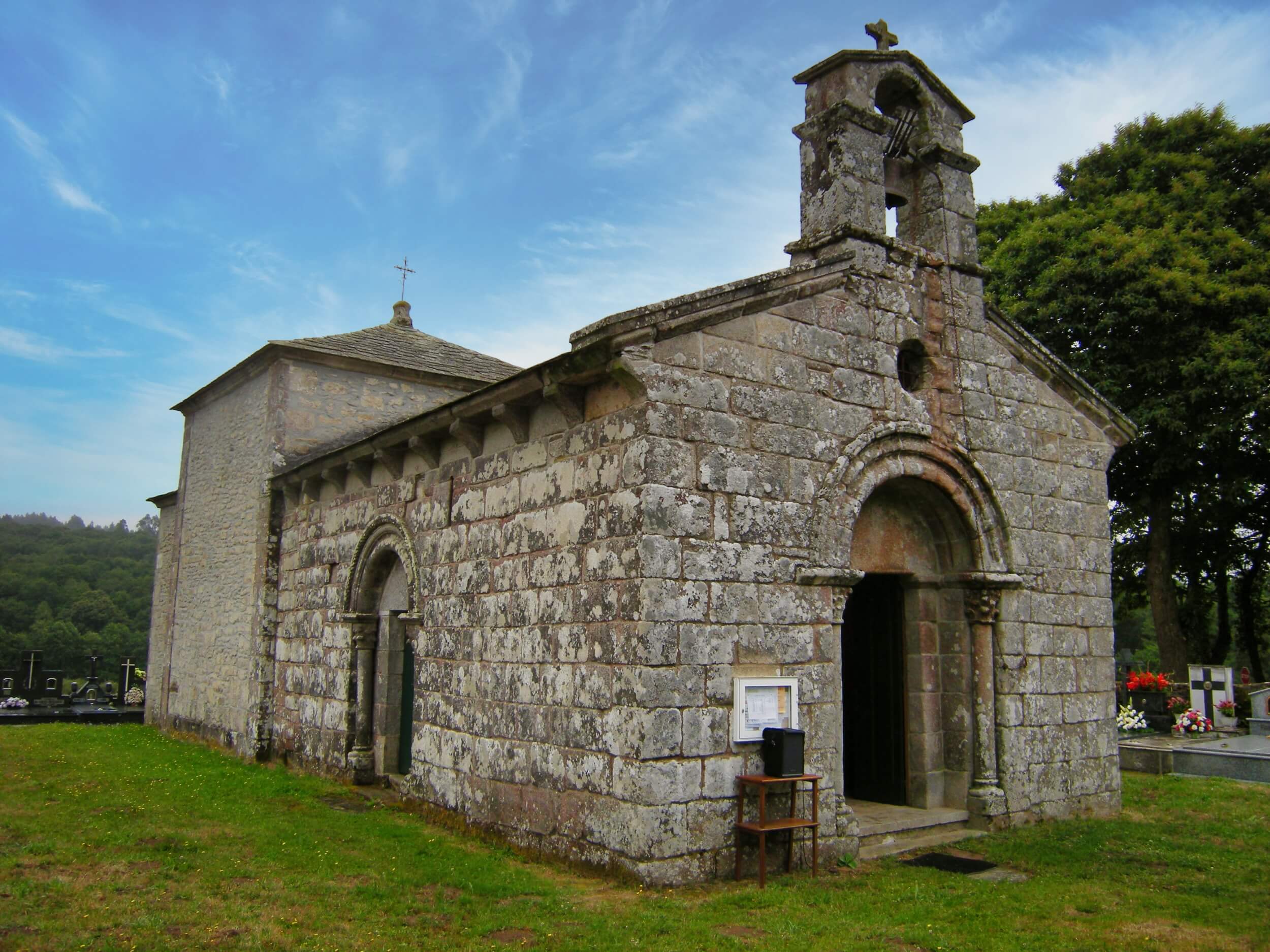 Petite église rurale romane en pierre, dotée de portes voûtées et d'un simple pignon à cloche, nichée au cœur d'un cimetière verdoyant parsemé de pierres tombales. L'église se détache sous un ciel bleu, avec des arbres et des collines en arrière-plan.