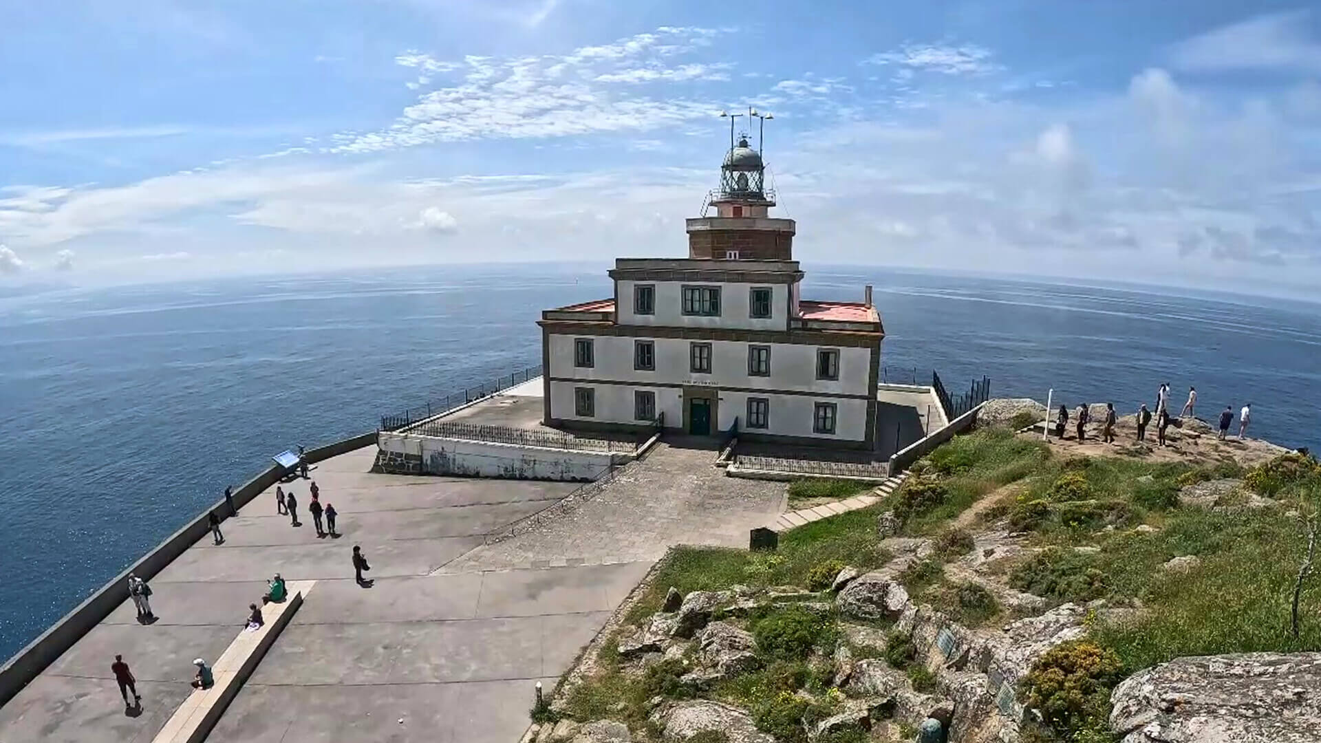 Au premier plan : une route goudronnée longeant une colline verdoyante et rocailleuse mène à un grand phare. Le phare trône au sommet d'un bâtiment rectangulaire de trois étages. À l'arrière-plan : l'océan Atlantique s'étend à perte de vue.